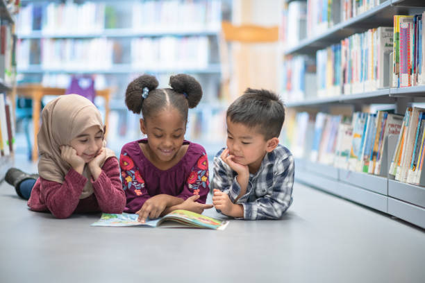 Friends who read together, stays together An adorable multi ethnic trio of preschool children and lying on the floor at their school's library as they share a book together. laying on belly stock pictures, royalty-free photos & images