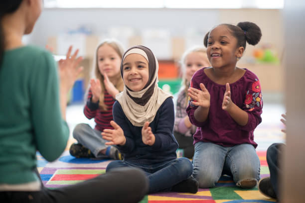 If you're happy and you know it, clap your hands! An adorable multi ethnic preschool children are singing and clapping their hands as their teacher is seated in front of them. community center stock pictures, royalty-free photos & images