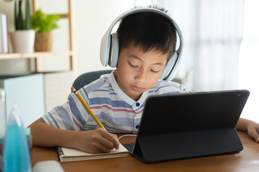 Asian boy learning online via internet with a tutor on a tablet digital with headphone, Asia child is studying while sitting in the living room at home. Concept of online learning at home