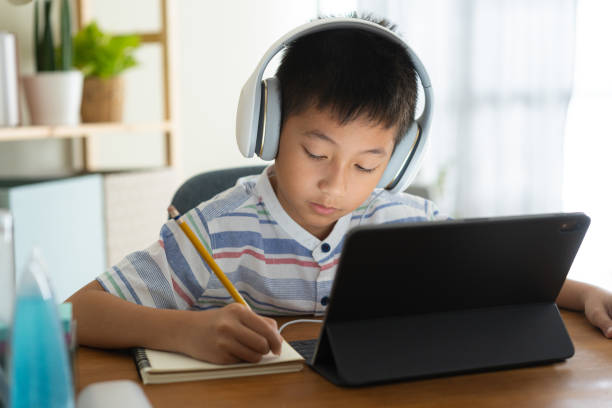 Asian boy learning online via internet with a tutor on a tablet digital with headphone, Asia child is studying while sitting in the living room at home. Concept of online learning at home stock photo