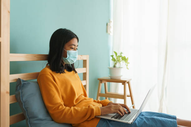 la donna asiatica indossa una maschera medica mentre è seduta a imparare online con il computer portatile a casa, concept apprendimento online a casa - home interior sparse bedroom inside of foto e immagini stock