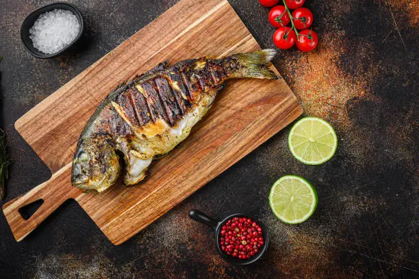 Photo of Grilled sea bream or dorado raw fish on chopping  board with ingredients over old metall dark background, top view with space for text.
