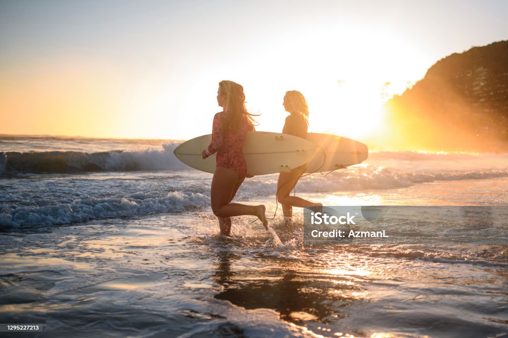 Young Female Surfers Running into Water at Burleigh Heads Full length side view with lens flare of women in teens and 20s wearing wetsuits and running into water with their boards at sunrise. Athlete Stock Photo