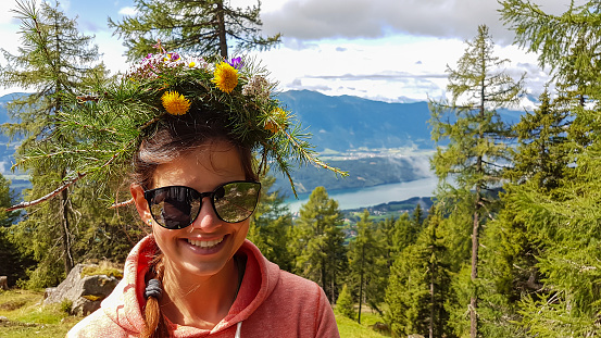 A woman in orange hoodie wearing a floral crown made of wild flowers and larch branches. Lush green pasture and dense forest. Many mountains poking above the clouds. Millstaettersee lake in the back.