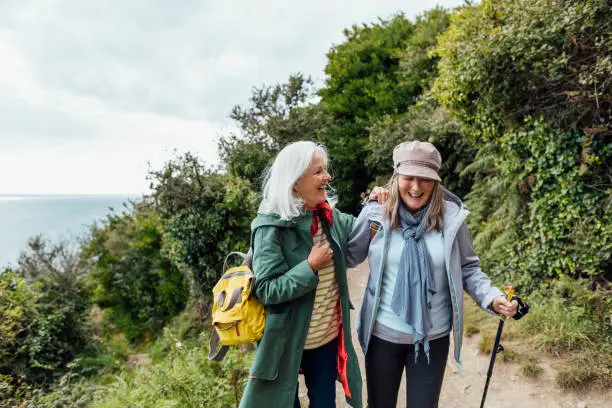 Female friends on a hike in Cornwall, they are walking on a coastal path, laughing while they have their arms around each other. The sea can be seen in the distance.