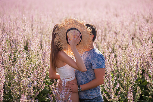 A happy couple in love hid behind a straw hat, kissing at sunset near a blooming sage field. A young man passionately embraces, holds the hands of a beautiful girl. Lovers weekend travel. Copy space