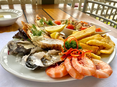 High angle closeup view of a delicious Seafood Platter in a Restaurant with fresh cooked Australian King prawns, fresh natural oysters in the half shell, half a cooked crayfish, octopus salad, lemon wedges, hot potato chips and a red capsicum strips and parsley garnish on a round white ceramic plate