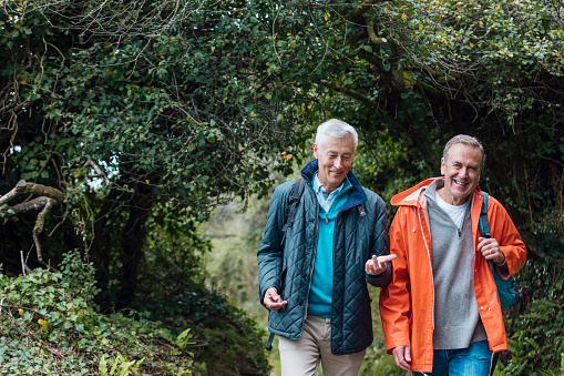 A senior and mature man walking on a coastal path through a tree area in Cornwall. They are talking to each other while they walk.