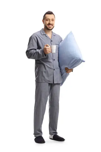 Full length portrait of a man in pajamas holding a pillow and a cup isolated on white background