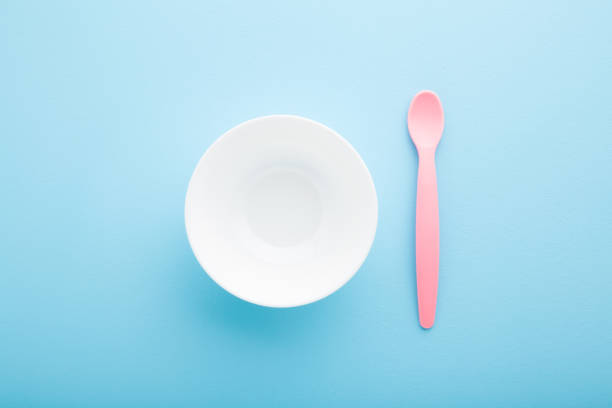 White bowl and pink plastic spoon on light blue table background. Pastel color. Closeup. Baby dishes. Top down view.