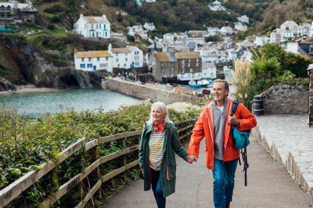 A Weekend Getaway A senior man and his wife holding hands walking up a hill on a footpath looking away from the camera at the view. The fishing village of Polperro is behind them. cornwall england stock pictures, royalty-free photos & images