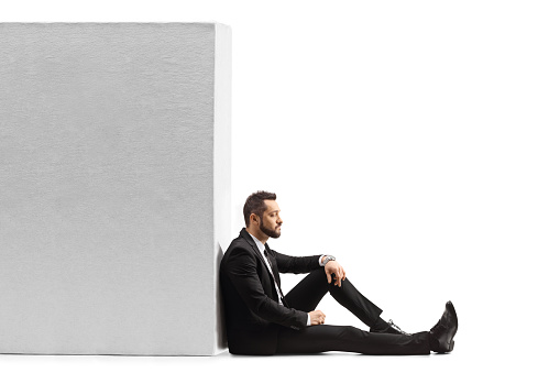 Pensive businessman sitting on the floor and leaning on a wall isolated on white background