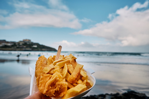POV of a hand holding a carton of Fish and Chips at Towan Beach, Newquay, Cornwall on an Autumn day.