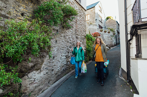 Two mature female friends walking downhill through a street in Polperro, Cornwall. They are carrying and pulling luggage, ready for a staycation in the fishing village. They are looking at the camera and smiling.