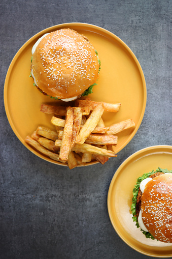 Stock photo showing elevated view of homemade chicken burgers served in sesame seed bread buns with melted cheddar cheese, tomato ketchup and lettuce served with chips on yellow plates.