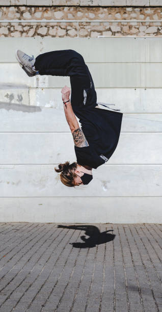 istock A young man wearing a mask somersaults backwards. Backflip. Float upside down with a concrete wall in the background. He wears a ponytail and wears black sportswear. Jump 1295312651
