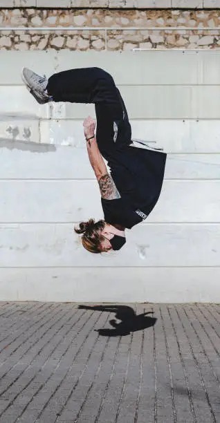 A young man wearing a mask somersaults backwards. Backflip. Float upside down with a concrete wall in the background. He wears a ponytail and wears black sportswear. Jump