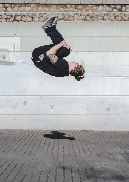 A young man wearing a mask somersaults backwards. Backflip. Concrete wall in the background. He wears a ponytail and wears black sportswear. Jump