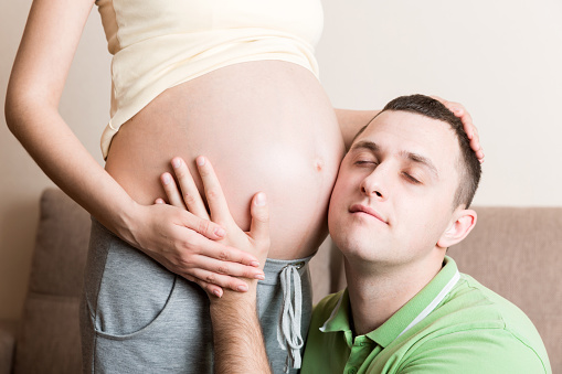 Young bearded man putting his ear close to his pregnant wifes abdomen and Listening it.