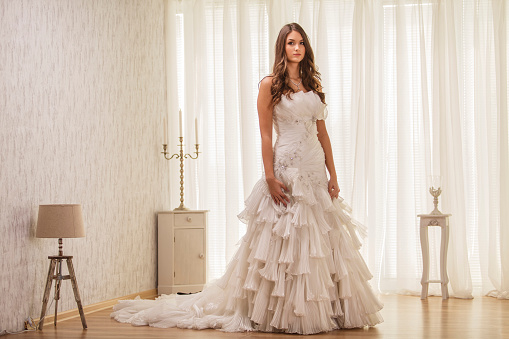 Wide shot of beautiful young bride in a elegant, riffled wedding dress standing in a room and confidently looking at camera.