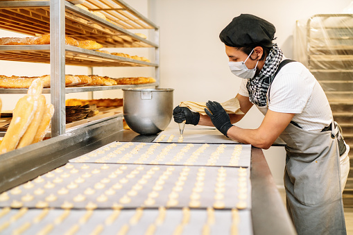 close-up of a pastry chef with confectionary bag squeezing cream in the pastry shop or bakery with gloves and face mask due to the 2020 covid19 coronavirus pandemic