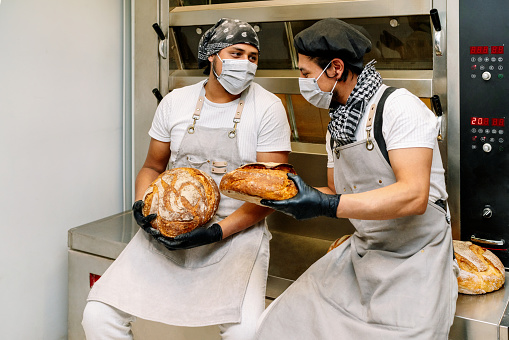 two happy latino bakers showing how they made a loaf of wheat bread in the bread oven with gloves on their hands and masks on their faces because of the 2020 covid19 coronavirus pandemic