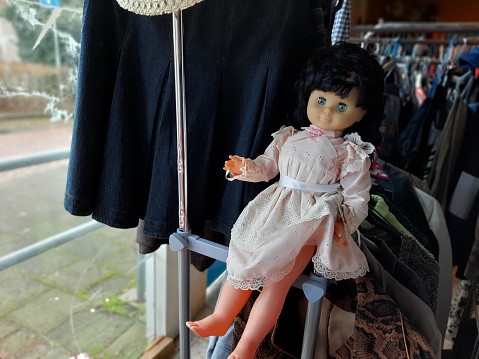 Doll at a garage sale or flea market in a pink dress with long brown hair and bleu eyes sitting on a cloth rack wit second hand cloths.