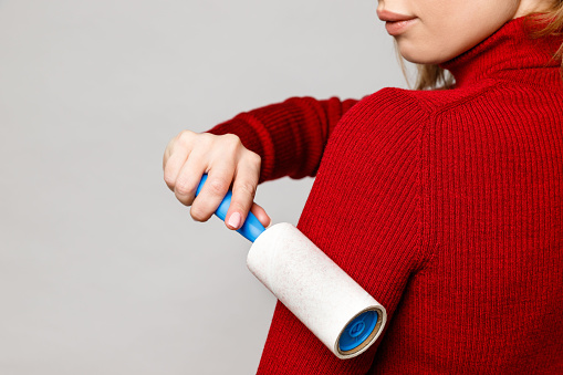 Woman hand using a sticky roller to clean fabrics - red woolen turtleneck from dust, hair, lint and fluff, front view, close up. Grey background.