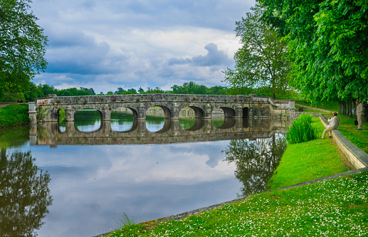 Chambord, France-May 26, 2014-A tourist sits on a low wall to take a photograph of the ancient arched stone bridge while his companion waits under the overhanging trees.