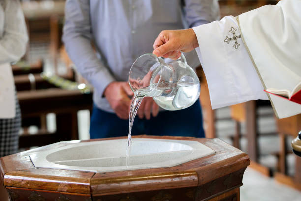 priest pouring holy water into the baptismal font, moments before a child receives the sacrament of baptism - batismo imagens e fotografias de stock