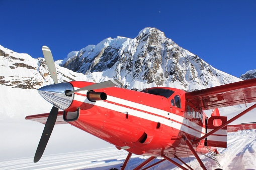 A crescent moon rises in a crystal clear, blue sky as an airplane sits at Ruth Glacier near Denali in the Alaska Range.