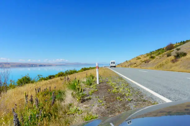 On highway around Lake Pukaki reflection in car bonnet of road ahead in view of turquoise water and blue sky.