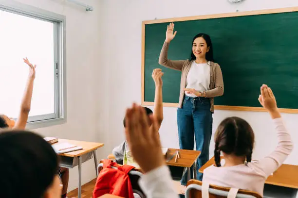 Photo of Asian school teacher with students raising hands. Young woman working in school with arm raised, school children putting their hands up to answer question, enthusiasm, eager, enjoyment