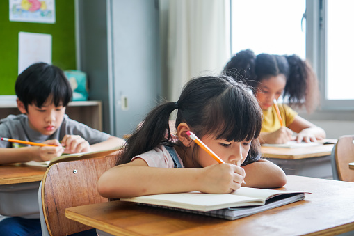 School girl sitting in school writing in book with pencil, studying, education, learning. Asian children in the class. Student diversity.