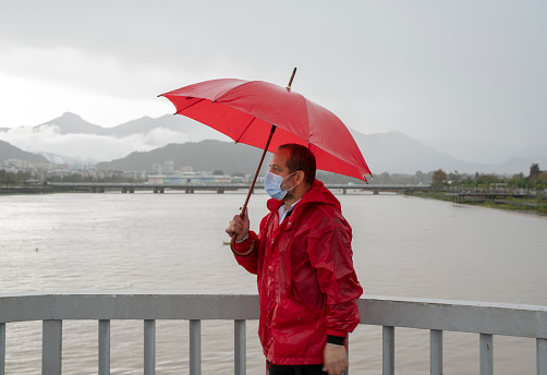 Mature man wearing red raincoat standing on the beach in rain with a red umbrella