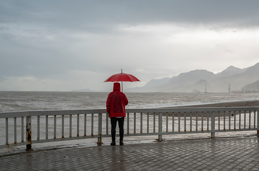Mature man wearing red raincoat standing on the beach in rain with a red umbrella