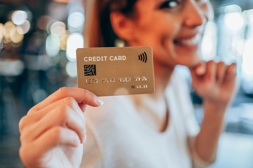Beautiful young female shopper holding a credit card - focus on foreground. Woman showing gold colored credit card against the camera.