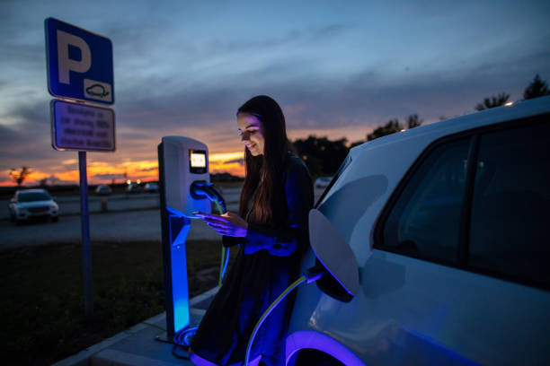 mujer cargando su coche eléctrico en la gasolinera por la noche. mujer usando el teléfono móvil mientras espera a que el coche eléctrico se cargue en el estacionamiento por la noche - urban scene women adventure city fotografías e imágenes de stock