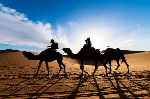 Photo of Stunning view of the silhouette of two people riding camels on the sand dunes in Merzouga, Morocco.