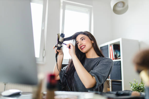 femme regardant l’ordinateur et utilisant son fer de curling tout en s’asseyant à la table d’habillage. la femme fait ses cheveux en regardant un tutoriel sur internet au moment de la pandémie de virus corona. - stage makeup white caucasian fashion photos et images de collection