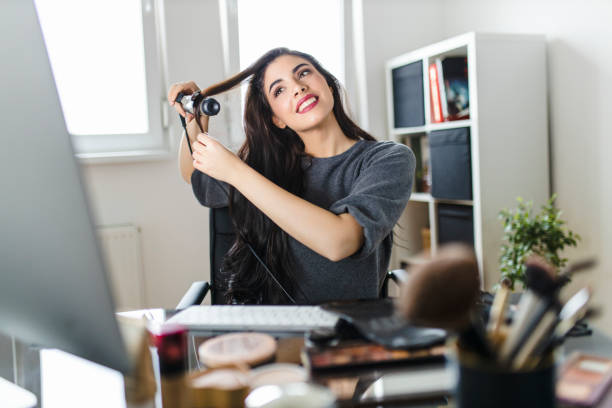 femme regardant l’ordinateur et utilisant son fer de curling tout en s’asseyant à la table d’habillage. la femme fait ses cheveux en regardant un tutoriel sur internet au moment de la pandémie de virus corona. - stage makeup white caucasian fashion photos et images de collection