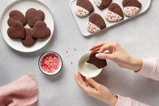 female hands dip chocolate cookie heart into melted white chocolate. baking with love for Valentines day. flat lay. Gray background