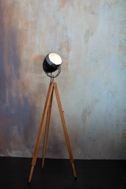 Stylish lamp on tripod in room. Tripod Light projector. Decorative light projector stands against a gray concrete wall. Loft living room interior. Floor lamp on wooden tripod. loft style floor lamp