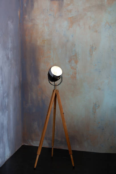 istock Stylish lamp on tripod in room. Tripod Light projector. Decorative light projector stands against a gray concrete wall. Loft living room interior. Floor lamp on wooden tripod. loft style floor lamp 1295465924