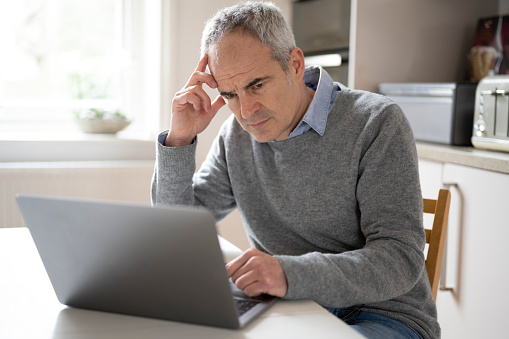 Mature caucasian man in his 50s using laptop looking concerned, puzzled and confused. He is seated at table in domestic kitchen. Cybe security problems.