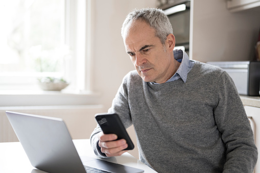 Mature caucasian man in his 50s using smart phone laptop looking concerned, puzzled and confused. He is seated at table in domestic kitchen. Cyber security issues.