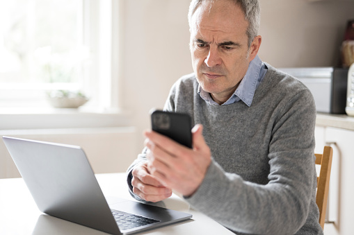 Mature caucasian man in his 50s using smart phone laptop looking concerned, puzzled and confused. He is seated at table in domestic kitchen. Cyber security issues.