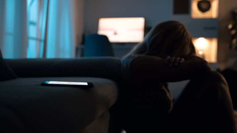 Depressed woman sitting on floor at night with mobile phone on sofa