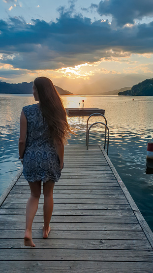 A woman in a blue dress walks along a wooden pier of Millstaettersee lake and enjoys the sunset. The sun is setting behind high Alps. Calm surface of the lake reflects the orange sky and the mountains