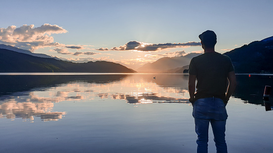 A man in a full cup standing at the shore of Millstaettersee and enjoying the sunset. The sun sets behind high Alps. Calm surface of the lake reflects the orange sky and the mountains. Meditation